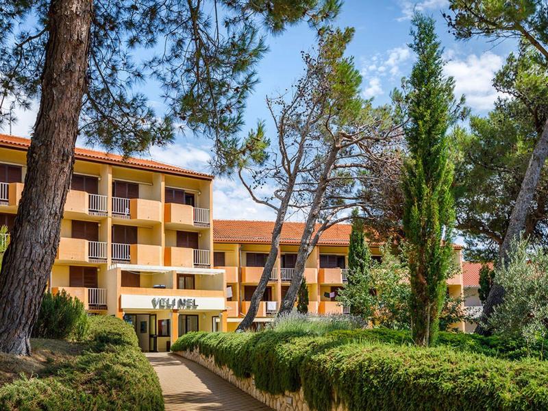 Mehrstöckiges gelb-oranges Gebäude umgeben von hohen Bäumen und grünen Hecken unter blauem Himmel.