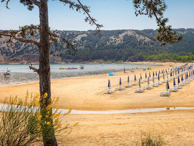 Breiter Sandstrand mit Reihen geschlossener blauer Sonnenschirme und Bergen im Hintergrund.