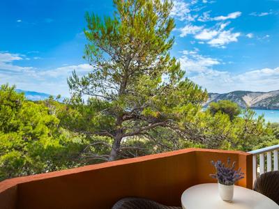 Balkon mit weißem Tisch, Vase und Lavendel, Blick auf grüne Bäume und blaues Meer unter blauem Himmel.