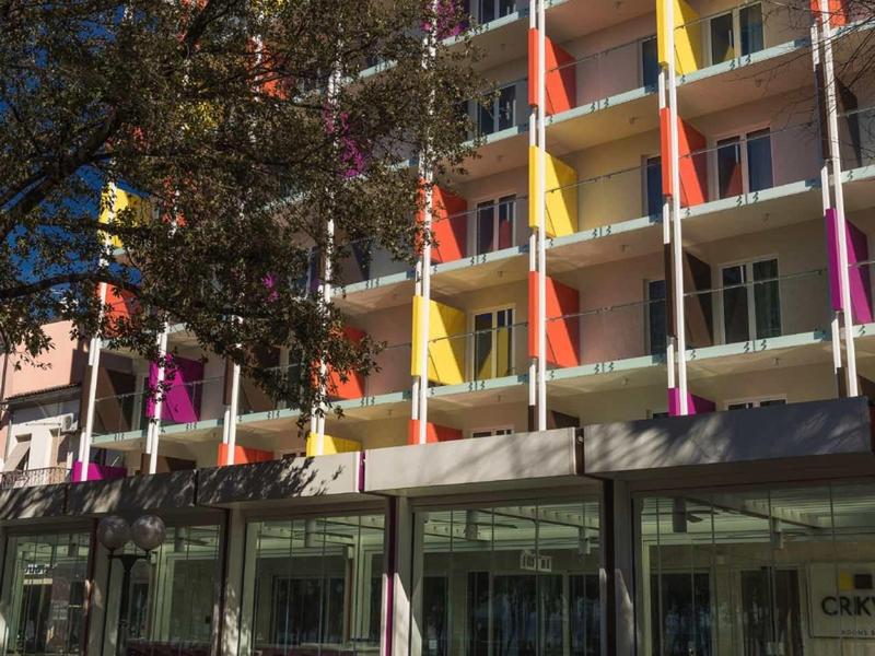 Multi-story hotel building with colorful balconies and street café in front on a sunny day.