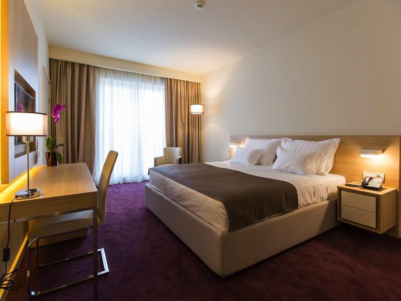 Modern hotel room with double bed, desk, chair, and window with curtains.