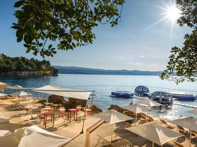 Sonnige Terrasse mit Sitzplätzen und Schirmen am ruhigen Seeufer mit Booten und Hügeln im Hintergrund.