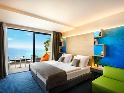 Habitación de hotel moderna con cama doble, sofá verde y vista al mar a través de puerta de cristal grande