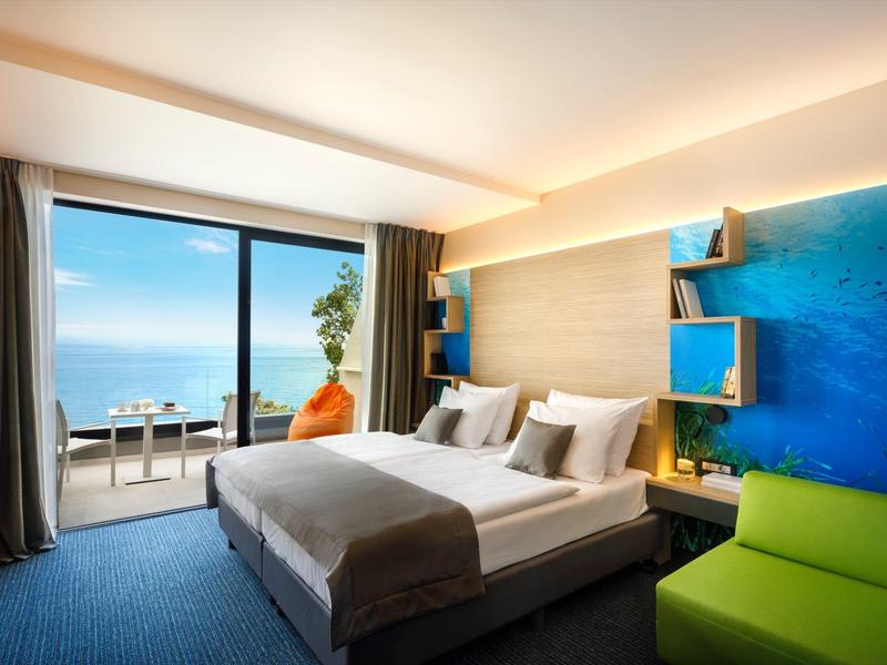 Modern hotel room with sea view balcony, comfortable bed, and vibrant blue and green decor.