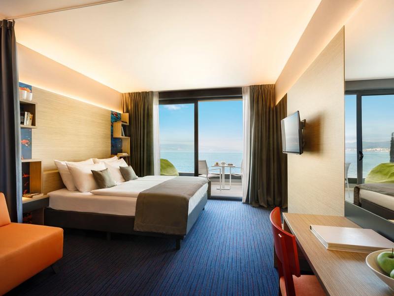 Modern hotel room with large bed, blue carpet, orange chair and sea view through window.