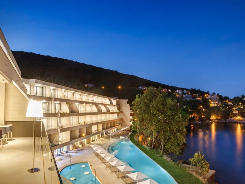 Modern hotel with a pool overlooking a calm lake at twilight with lights reflecting in the water.