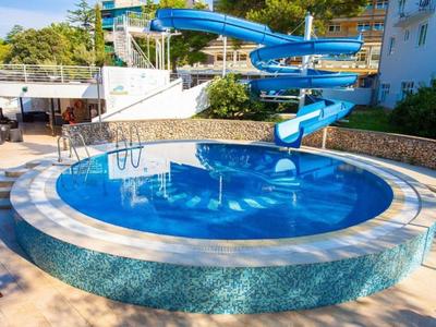 Piscina redonda al aire libre con tobogán azul y escaleras, rodeada de árboles y edificios.