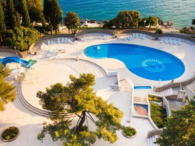 Blick auf einen Poolbereich mit Wasserrutsche und Liegestühlen, umgeben von Bäumen und Meeresblick.