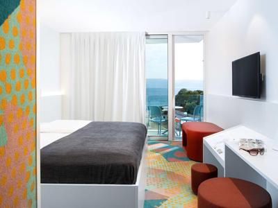 Lichte hotelkamer met tweepersoonsbed, aan de wand gemonteerde tv en balkon met uitzicht op zee.