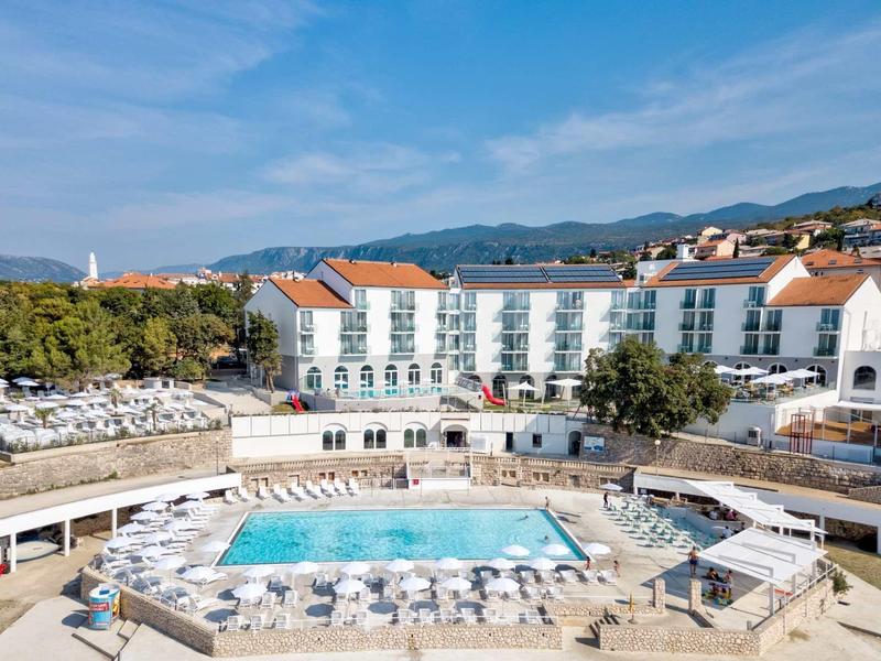 Hotel with large outdoor pool and terrace filled with sun loungers against a mountain backdrop.