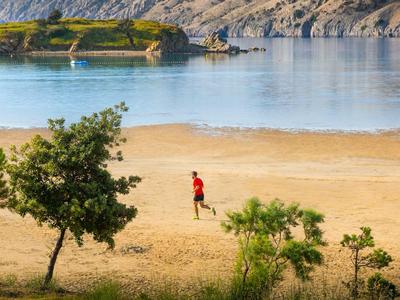 Person wandert am sandigen Ufer mit ruhigem Wasser und Felsen im Hintergrund.