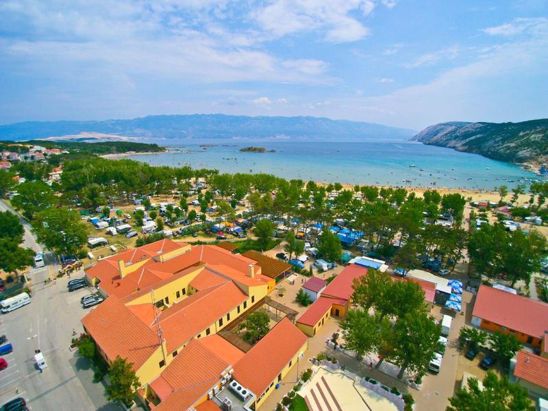 Aerial view of a hotel with red roofs by the coast with beach and mountains in the background.