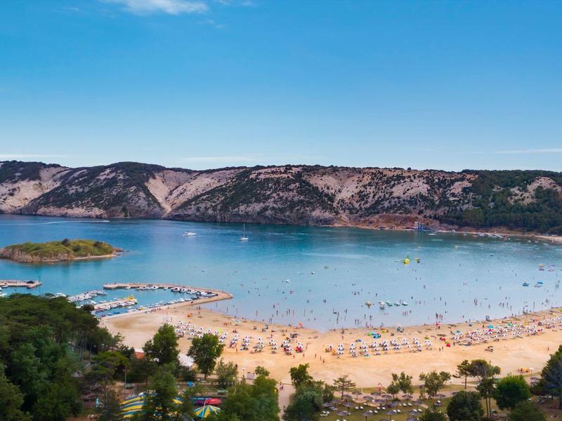 Beach with golden sand and clear blue water in front of rocky hills and a blue sky.