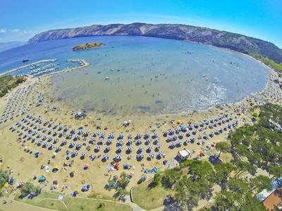 Vista panorámica de una playa de arena concurrida con sombrillas frente a una bahía tranquila y colinas al fondo.