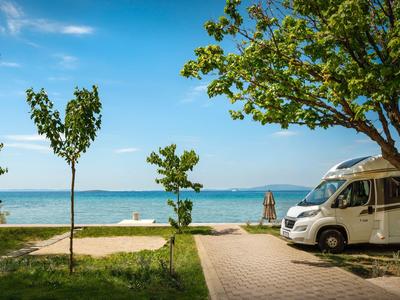 Autocaravana aparcada junto a un árbol con vistas a un lago y cielo azul.