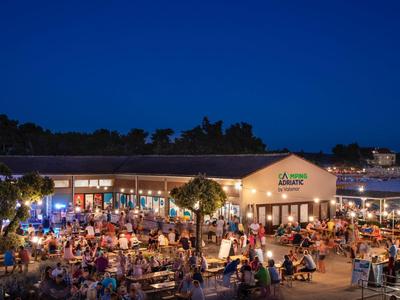 Área exterior concurrida de un restaurante junto al agua por la noche con mucha gente y luces.