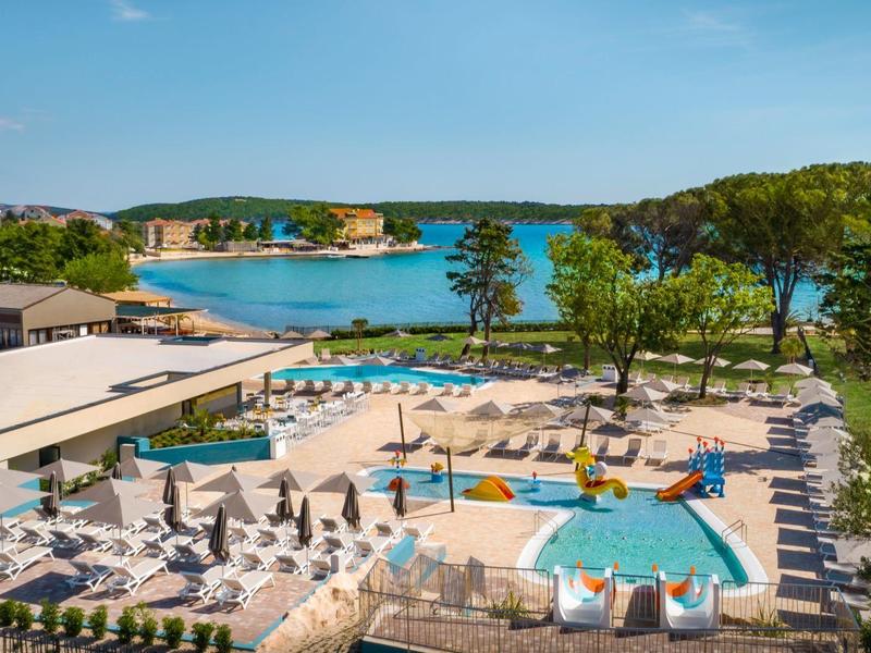 Area piscina moderna con lettini, ombrelloni e vista sul mare blu e sul paesaggio verde.