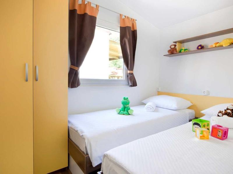 Bright bedroom with two single beds, yellow wardrobe, and window with brown curtains.