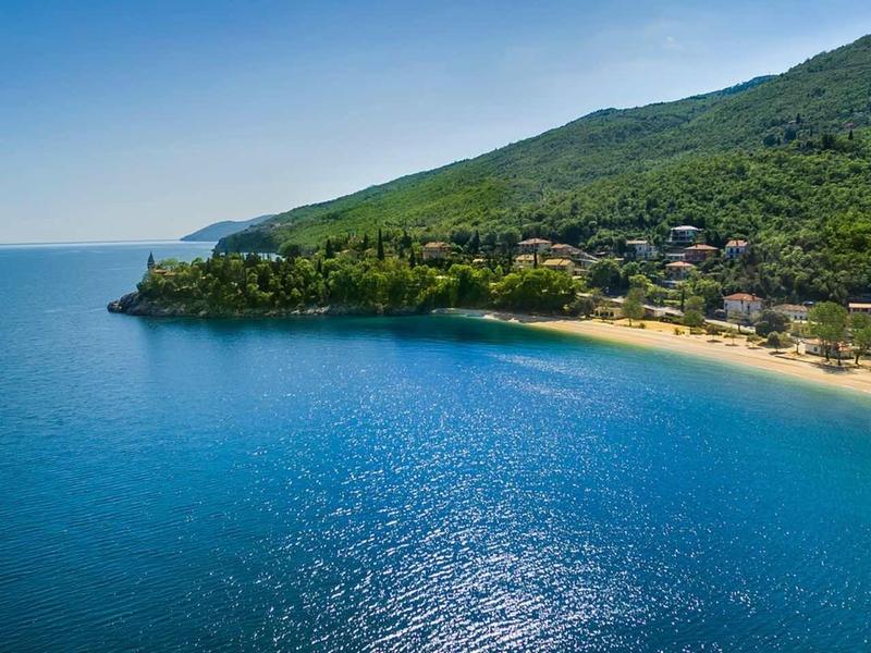 Coastal landscape with blue sea, sandy beach, and forested hills under clear sky.