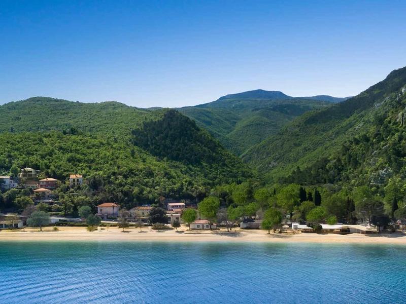 A tranquil sandy beach in front of a forested, hilly coastal village.