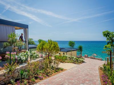Gepflasterter Weg führt durch blühenden Garten mit Blick auf das Meer unter blauem Himmel.