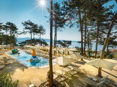 Hotelpoolbereich mit Liegestühlen und Sonnenschirmen, umgeben von Pinien und Blick aufs Meer.