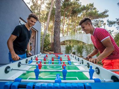 Dva mladí muži hrají stolní fotbal venku u budovy s stromy v pozadí.