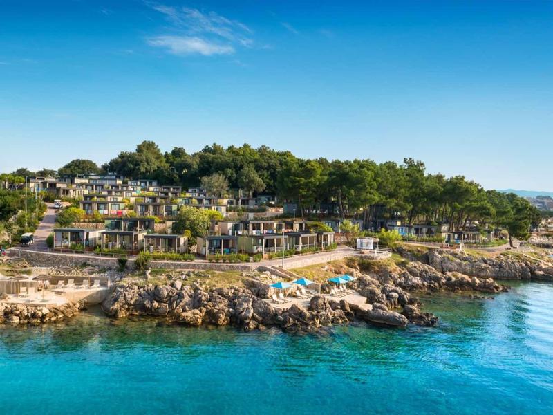 Moderní rekreační domy na skalnatém poloostrově s výhledem na modré moře a zalesněné kopce.