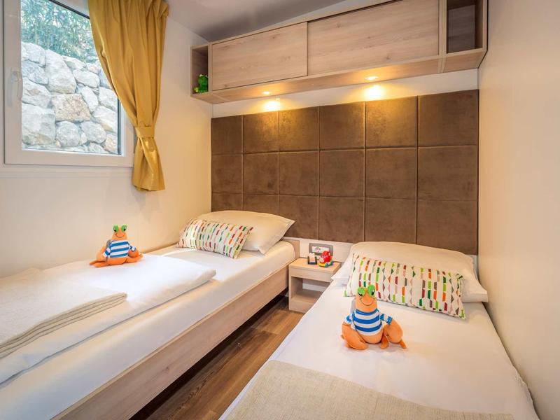 Dvoulůžkový pokoj s dřevěnými postelemi, světly nahoře a oknem se žlutou záclonou.