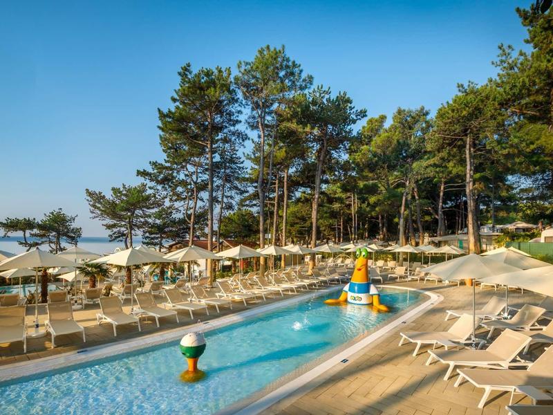 Bazénová oblast s lehátky, slunečníky, stromy a modrou oblohou v pozadí