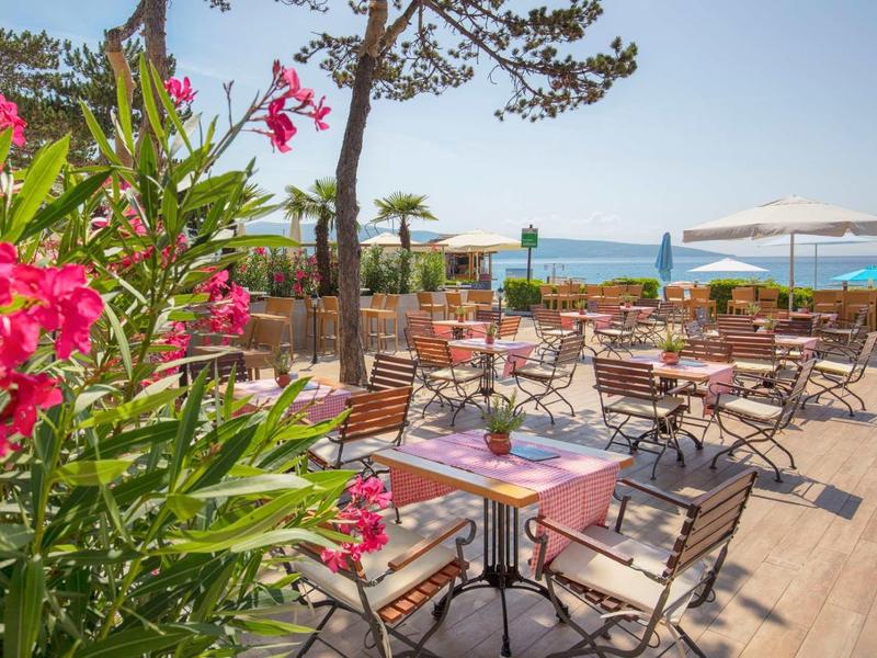 Terasa s stoly, židlemi a květinami s výhledem na moře za slunečného dne.