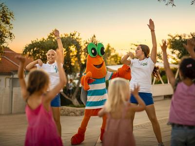 Children dancing with two entertainers and a person in a colorful fish costume outdoors.
