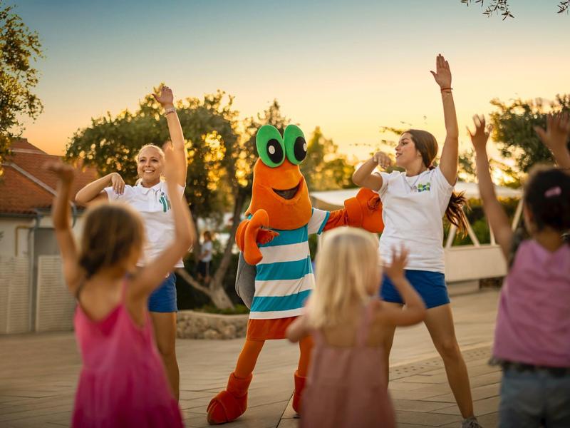 Children dancing with two entertainers and a person in a colorful fish costume outdoors.