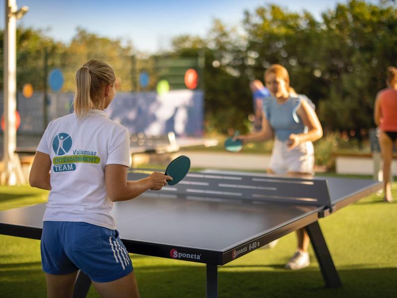Person playing table tennis outdoors on a sports field with green areas in sunlight.