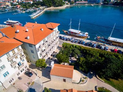 Uitzicht op meerlaagse hotel met rode daken aan haven met boten en blauw water.