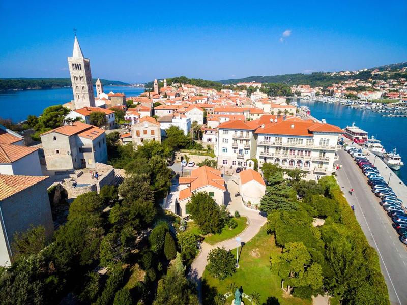 Vista de una ciudad costera con puerto, techos rojos y jardines verdes bajo cielo azul.