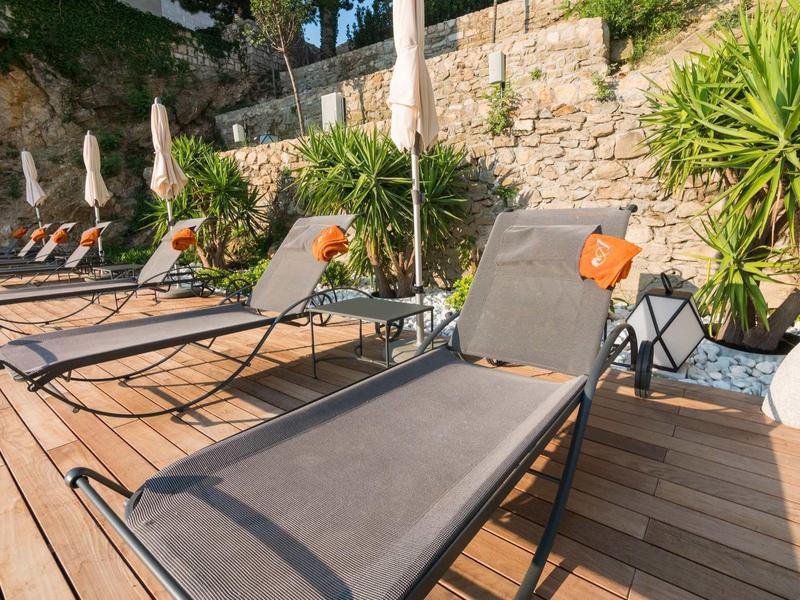 Varias tumbonas en una terraza de madera junto a un muro de piedra y plantas en el jardín del hotel.
