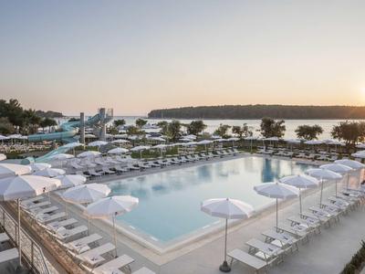 Großer Pool mit weißen Liegen und Schirmen, Palmen und Meer im Hintergrund bei Sonnenuntergang.