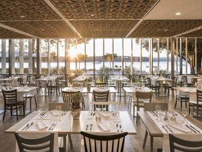 Velká restaurace s mnoha prostřenými stoly a velkými okny s výhledem na vodu při západu slunce.