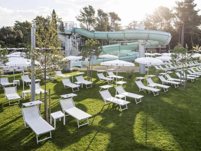 Wiele białych leżaków i parasoli na zielonej trawie przed zewnętrzną zjeżdżalnią wodną.