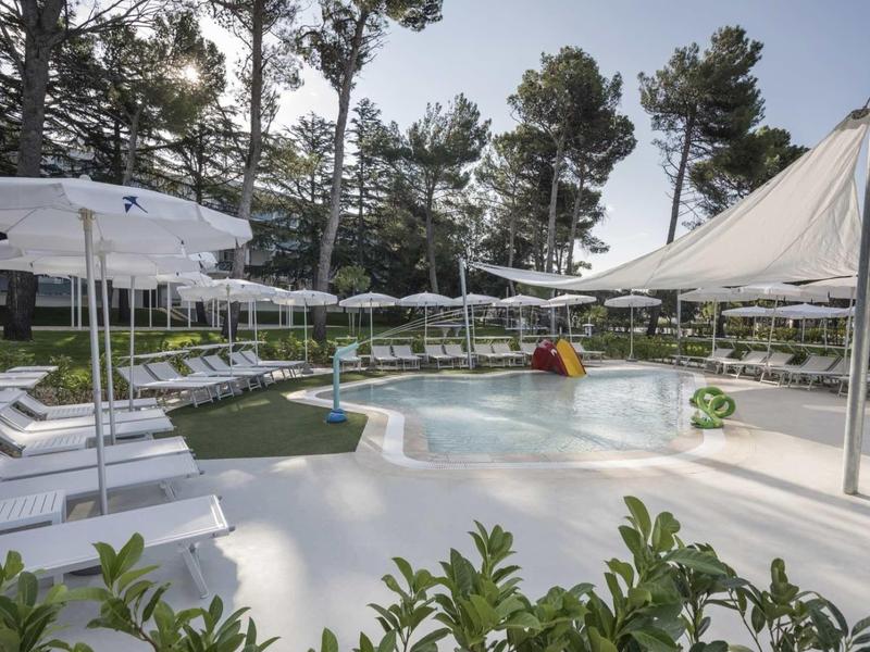 Basen dla dzieci z parasolami i leżakami w hotelu z drzewami w tle.