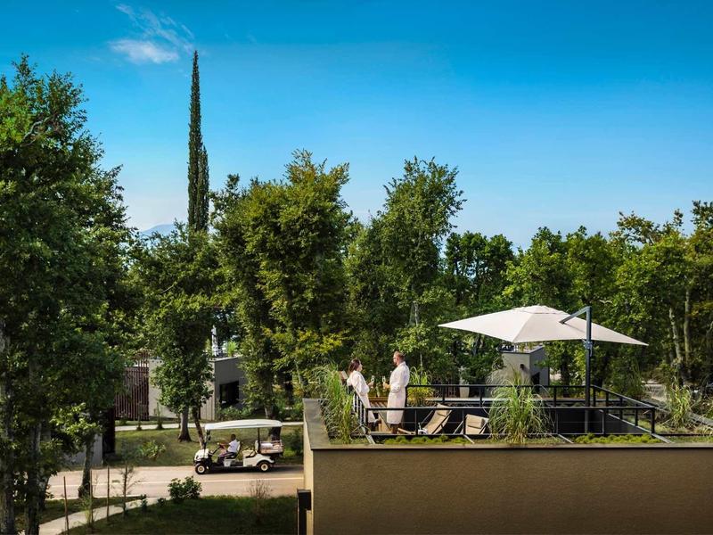 Terrace with parasol and guests, surrounded by green trees under clear sky.