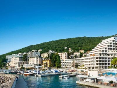 Hotel complex with multiple buildings on the coast and marina in front of a forested hill.