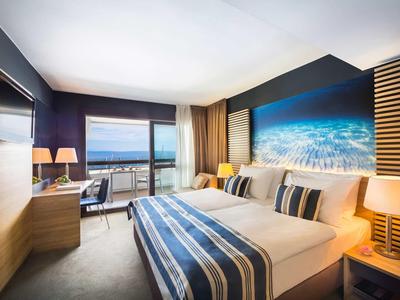 Modern hotel room with double bed, sea view, and seating area by floor-to-ceiling windows.