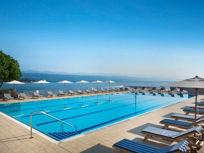 Large outdoor pool with loungers and umbrellas, overlooking the sea under clear skies.