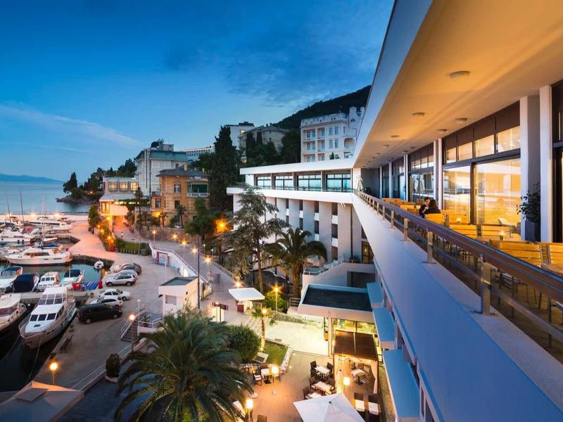 View of a marina at sunset with a modern hotel and terrace.