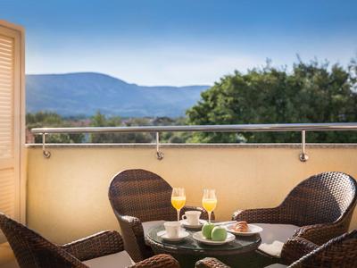 Balcón con sillas de mimbre y mesa, vista a montañas y cielo azul.