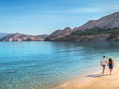 Una pareja camina por una playa tranquila con agua azul clara y montañas al fondo.