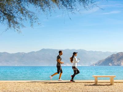 Zwei Personen joggen am Sandstrand mit Meer und Bergen im Hintergrund unter blauem Himmel.