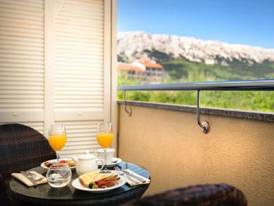 Kleiner Balkon mit Rundtisch, zwei Stühlen, Frühstück und Blick auf Hügel mit Häusern.